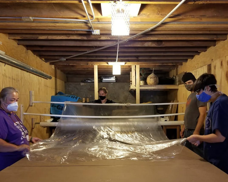 Four people at Future Forge Makerspace in New Mexico stand at a table rolling out plastic for making isolation gowns.