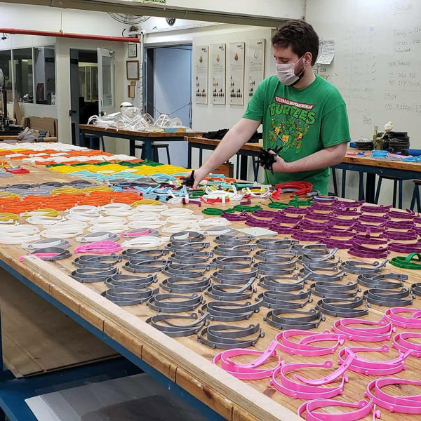 Person in Teenage Mutant Ninja Turtles tshirt lays out dozens of 3d printed face shield parts in a variety of colors.